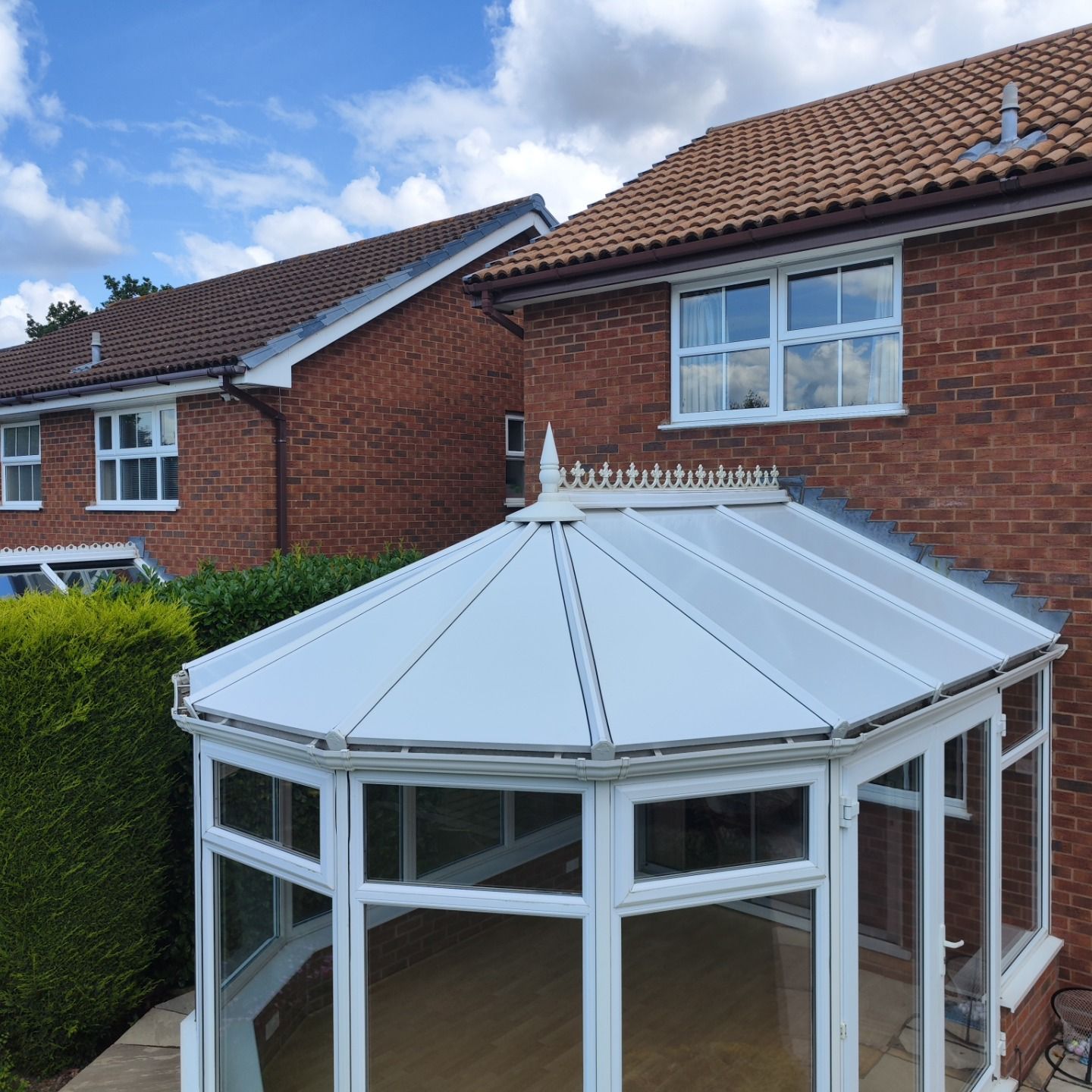 Insulated conservatory roof with a white finish, featuring a pointed apex and multiple glass panels, adjacent to a brick house and greenery, showcasing year-round comfort solutions by Rosy Roof.